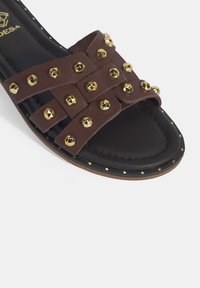 Bruine leren slipper met gouden studs, gekruiste banden, een platte zool en decoratieve stiksels langs de randen.