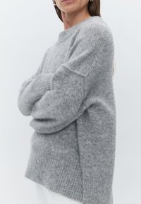 Sweater cinza, tricotado, com um corte solto, bainha canelada e mangas largas. Textura suave com uma aparência ligeiramente felpuda.