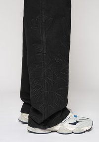 Pantalon en denim noir avec un motif brodé de fleurs en relief sur la jambe gauche ; assorti à des baskets volumineuses blanches et grises.