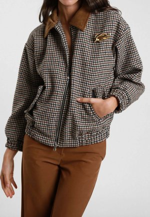 Giacca a chevron marrone e beige con colletto in corduroy, zip frontale e tasche laterali. Presenta un piccolo dettaglio metallico sul petto.