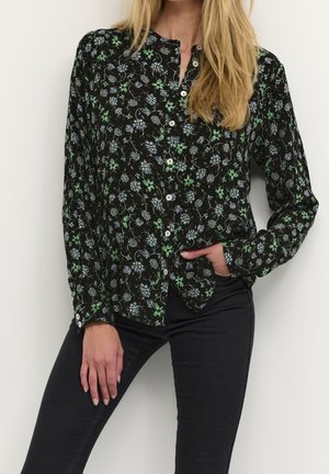 Femme portant un chemisier noir à manches longues avec un imprimé floral vert et blanc, associé à un pantalon noir ajusté, debout devant un fond uni.