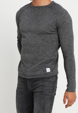 Mężczyzna w dopasowanym ciemnoszarym swetrze z długim rękawem i ciemnoszarych dżinsach, stojący z lekko wyprostowanym prawym ramieniem.