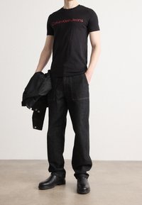 Černé bavlněné tričko s červeným nápisem "Calvin Klein Jeans", kombinované s tmavě modrými džínami, černými botami a černou bundou přehozenou přes ruku.