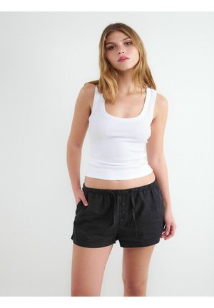 Młoda kobieta ubrana w biały top bez rękawów i czarne krótkie spodenki na guziki, stojąca z rękami w kieszeniach na tle jednolitego białego tła.