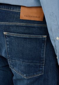 Zbliżenie na tylną kieszeń ciemnoniebieskich dżinsów z brązową skórzaną naszywką z napisem "FAÇONNABLE" oraz osobę ubrana w jasnoniebieską koszulę.