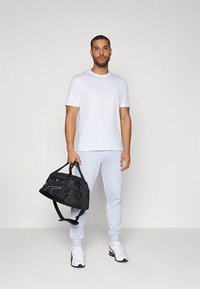 T-shirt branca de mangas curtas, calças de ginásio cinza claro, a transportar uma bolsa de ginásio preta. Ténis brancos com detalhes em vermelho e preto. Design e ajuste simples.