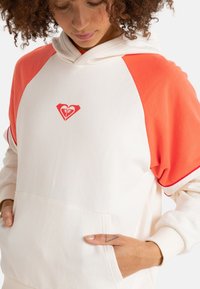 Hooded sweatshirt with a cream body and bright orange raglan sleeves. Features a front pocket and a small red heart logo on the chest.