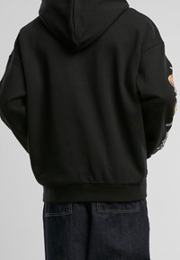 Osoba v černé mikině s barevnými vzory na pravém rukávu, v kombinaci s tmavě modrými džíny, otočená zády k fotoaparátu.