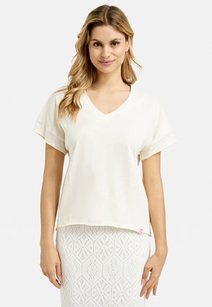 V-neck short-sleeve top in cream, made of lightweight fabric with a relaxed fit; tag visible at the hem, paired with a patterned white skirt.
