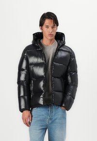 Black puffer jacket with a glossy finish, featuring a hood, zip closure, and horizontal quilted design. Worn over a gray sweater and blue jeans.