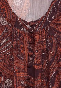 Primer plano de tela con un patrón de paisley en rojo oscuro y negro, que presenta pequeños botones cubiertos a lo largo de un escote plisado.
