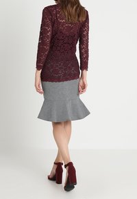Burgundy spetsblus med långa ärmar, lagd över en grå pennkjol; kjolen har en utvidgad fåll. Kombineras med öppna klackar.