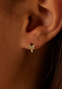 Boucle d'oreille en or avec un petit papillon orné de pierres précieuses transparentes sur l'oreille d'une personne, contre un fond sombre.