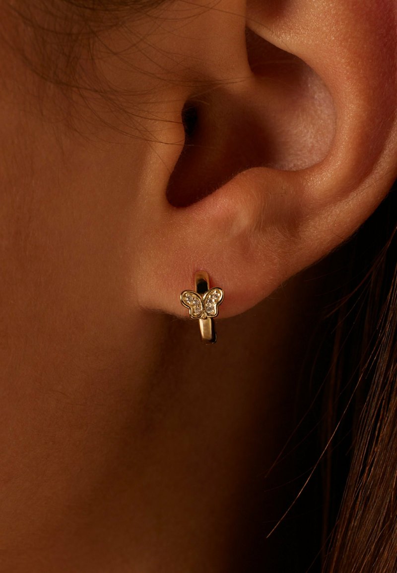 Boucle d'oreille en or avec un petit papillon orné de pierres précieuses transparentes sur l'oreille d'une personne, contre un fond sombre.