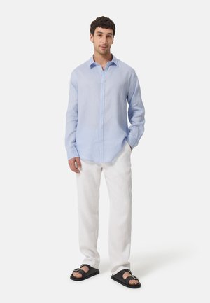 Camisa de lino de color azul claro con cuello y cierre de botones, combinada con pantalones de lino blancos y sandalias negras.
