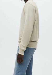 Uomo che indossa una felpa beige a maniche lunghe e jeans azzurri chiari, in piedi contro uno sfondo chiaro e uniforme, rivolto di lato.