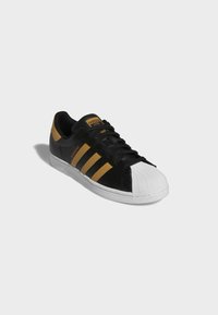 Black sneaker with three gold stripes, white rubber toe cap, and black laces. Features a branded tongue and textured leather upper.