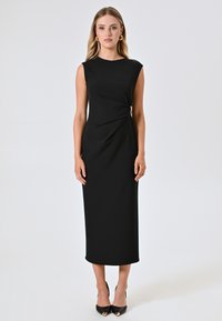 Robe midi sans manches noire avec un détail tordu asymétrique et des accents dorés. Tissu lisse, silhouette ajustée et design longueur cheville.