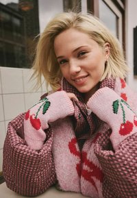 Rožinė megzta palaidinė su raudonų vyšnių raštais, dėvima po checkered pūstuoju rožiniu paltu, demonstruojanti tekstūruotą medžiagą ir kontrastingas spalvas.