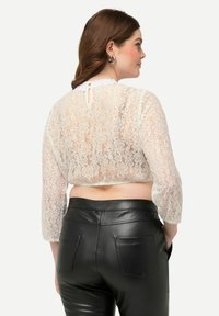 Top in pizzo bianco corto con maniche lunghe semi-trasparenti, caratterizzato da un colletto ondulato e un design a chiave sulla schiena, abbinato a pantaloni in pelle nera.