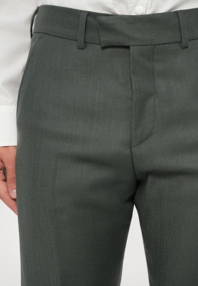 Pantaloni di un verde scuro con una texture e un taglio sartoriale, caratterizzati da un fronte piatto, passanti per la cintura e tasche laterali, abbinati a una camicia bianca.