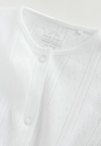 Weißes Baby-Cardigan mit feiner Stricktextur, versehen mit kleinen Diamantmustern und einem Druckknopfverschluss. Etiketten sind im inneren Halsausschnitt sichtbar.