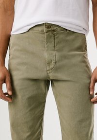Pessoa vestindo calças verde-oliva e uma camisa branca, de pé com as mãos relaxadas ao lado do corpo, em um fundo branco liso.