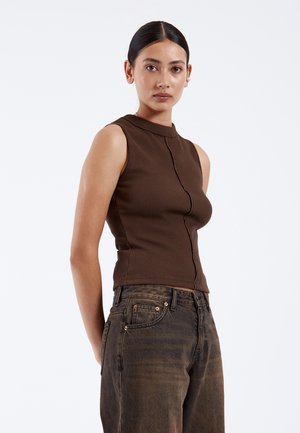 Joven mujer con cabello oscuro recogido en un moño bajo, lleva un top marrón sin mangas con textura y vaqueros negros descoloridos, de pie frente a un fondo blanco liso.