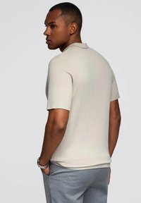 Polo de manga corta en color beige claro con textura acanalada y cuello, combinado con pantalones grises. Modelo visto desde la parte trasera, mostrando el ajuste.