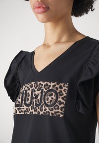 Top negro con mangas cortas y volantes, y escote en V. Presenta un panel de estampado de leopardo con "LIU JO" en letras con tachuelas. Tejido suave.