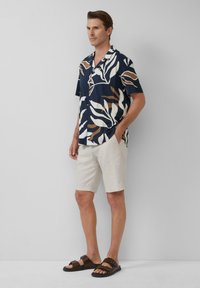 Mann in braun-weißer, blattmusterter, navyblauer Kurzarmbluse, hellbeigen Shorts und dunkelbraunen Sandalen steht vor einem schlichten Hintergrund.