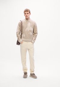 Young man standing with hands in pockets wearing beige patterned sweater, cream pants, taupe sneakers, and crossbody bag against white background.