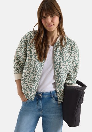 Kobieta z długimi brązowymi włosami, ubraną w zielono-beżową kurtkę w panterkę na białej koszuli i niebieskich dżinsach, trzymającą czarną pikowaną kurtkę.