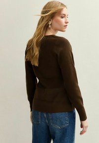 Maglione marrone a maglia con vita aderente e texture a coste, abbinato a jeans blu a vita alta. I capelli fluiscono delicatamente nella brezza.