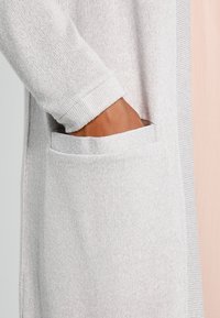 Světlé šedé pletené sako s žebrovanou strukturou, s viditelnou kapsou na levé straně a dlouhými rukávy.