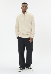 Béžový pulóver na zips s vysokým golierom, v kombinácii s voľnými čiernymi nohavicami a čierno-bielymi teniskami na jednoliatom pozadí.
