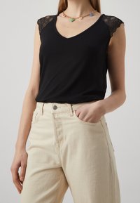 Sorte ærmeløs top med blonde detaljer ved skuldrene og V-udskæring. Sammen med høj talje beige bukser, der har en frontlomme.