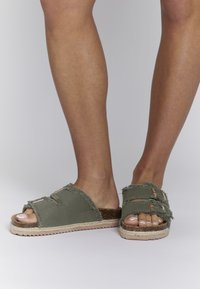 Green fabric slide sandals with frayed edges, cork footbed, and accent straps. Textured sole and open toe design for breathability.