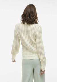 Pull en tricot de couleur crème clair avec un design texturé et à motif ouvert. Il a des manches longues et un ourlet côtelé. Complété par un pantalon à motifs pâles.
