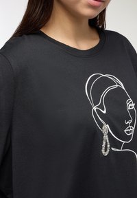 Czarna koszulka z białym rysunkiem twarzy, ozdobiona srebrną kryształową kolczykiem po lewej stronie.