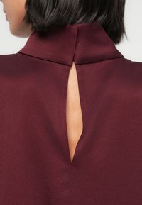 Burgundinpunainen paita korkealla kauluksella ja ainutlaatuisella takahalkiolla, jossa on sileä, teksturoitu kangas ja siisti, minimalistinen muotoilu.