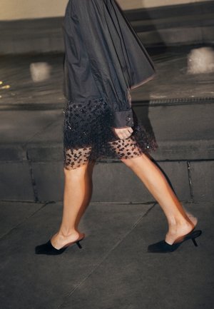 Chemise noire à boutons avec manches longues associée à une jupe noire transparente ornée d'un motif floral en perles ; escarpins noirs à bout ouvert.