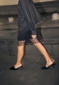 Chemise noire à boutons avec manches longues associée à une jupe noire transparente ornée d'un motif floral en perles ; escarpins noirs à bout ouvert.