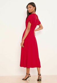 Červené midi šaty s krátkymi rukávmi, detailom v tvare kľúčovej dierky na chrbte a rozšírenou sukňou. Hladká látka, jednoduchý dizajn, v kombinácii s čiernymi sandálkami na vysokom podpätku.