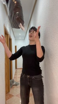 Persoon in een zwart shirt en spijkerbroek die een met plastic verpakt voorwerp omhoog gooit in een smalle, betegelde gang met witte muren.