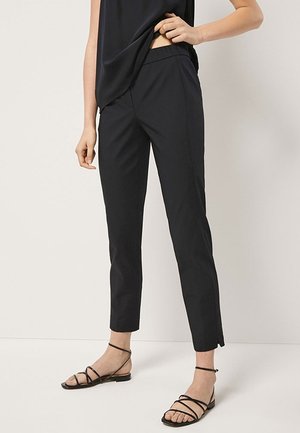 Femme portant un pantalon noir slim-fit jusqu'aux chevilles associé à des sandales plates noires à lanières, se tenant contre un fond clair uni.