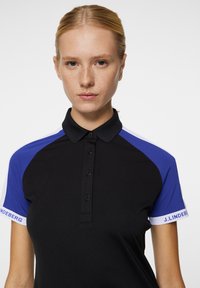 Svart polo skjorta med blåa accenter på axlarna, med krage, fyra knappar och märkt text på ärmen. Mjuk, åtsittande material.