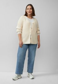 Cremfarbener Cardigan mit geripptem Textur, vier Knöpfen und tiefen Schultern, kombiniert mit hellblauen geraden Jeans und weißen Sneakers.