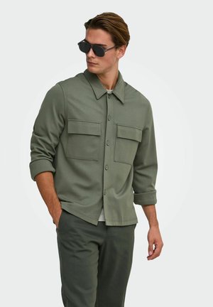 Homme portant une veste boutonnée vert foncé avec deux poches poitrine, un pantalon assorti et des lunettes de soleil noires, regardant sur le côté avec une main dans la poche.