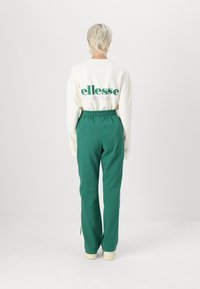 Kremveida džemperis ar zaļu "ellesse Ski Club" tekstu, apvienots ar augsto jostu zaļām sporta biksēm, kurām ir nelieli sānu iegriezumi apakšmalā.